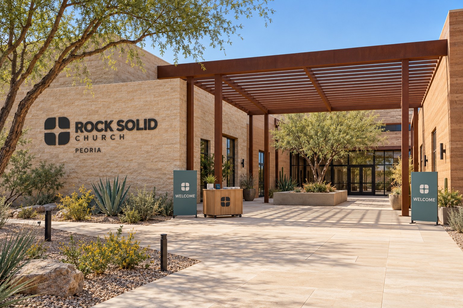 Peoria's warm masonry courtyard campus, with a rust-colored steel pergola, desert planting, and an empty Sunday welcome cart.